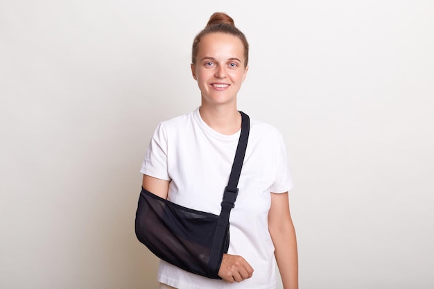 Portrait of positive satisfied woman with bun hairstyle wearing casual t shirt posing isolated over white background standing with hand in sling after accident