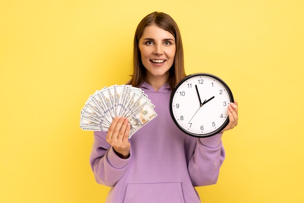 Photo portrait of positive smiling woman holding big fan of dollar banknotes and wall clock, time is money, expressing happiness, wearing purple hoodie. indoor studio shot isolated on yellow background.