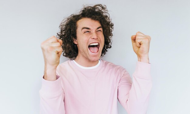 긍정적인 성공적인 잘생긴 젊은 남성의 초상화는 기쁨에 주먹을 꽉 쥐고 분홍색 스웨터를 입고 흰색 스튜디오 배경 위에 고립 된 행복으로 외치는 대로 입을 크게 벌립니다.