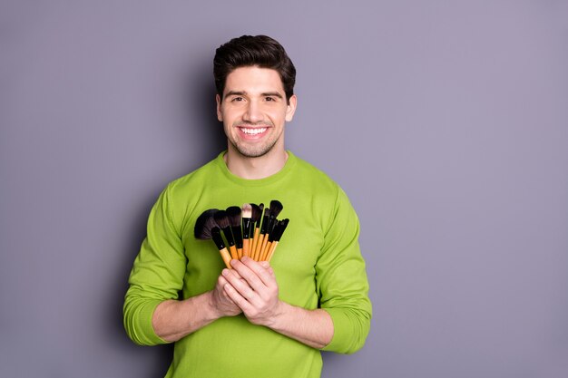 Portrait of positive successful man visagiste hold brushes to make maquillage for special evening events photo session wear green sweater isolated over grey color wall