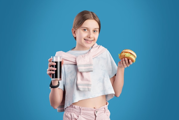 Portrait of positive teenage girl eating unhealthy hamburger and drinking cola