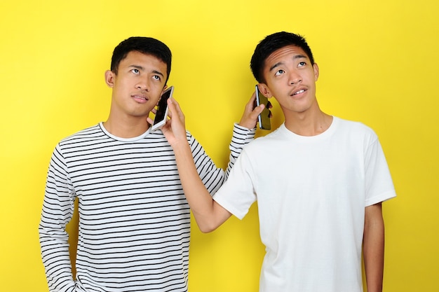 Portrait of positive two Asian men listen to each other's cellphones look at copy space isolated yellow background