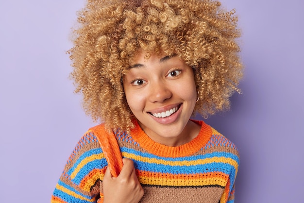 Portrait of positive woman with fair curly hair looks gladfully
at camera has good mood dressed in casual knitted colorful sweater
isolated over purple background happy people and emotions
concept