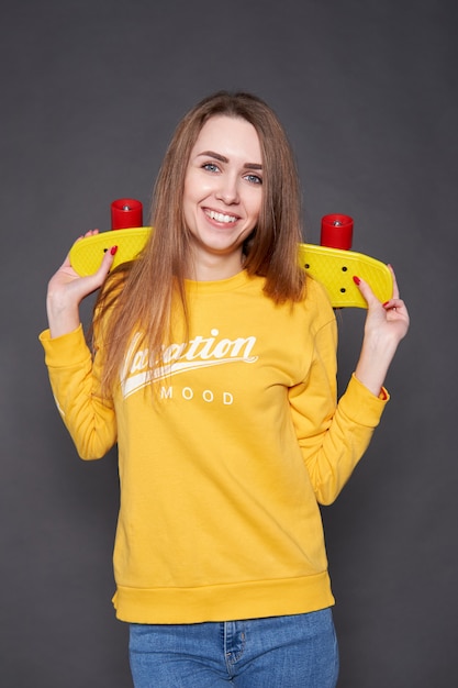 Portrait of positive young attractive girl wearing yellow blouse and blue jeans holding yellow skateboard.