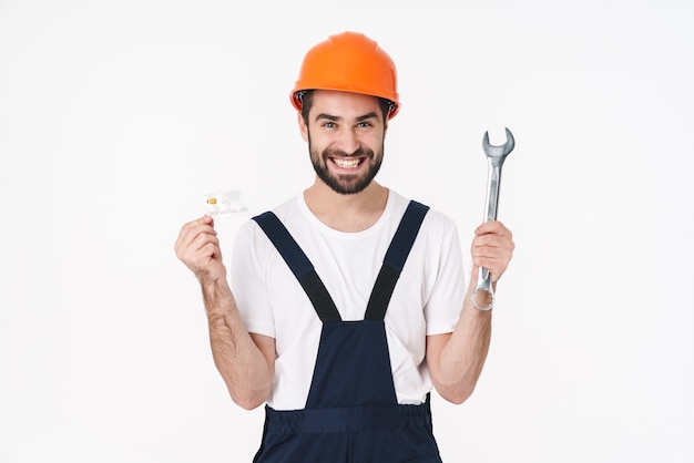 Portrait of positive young man builder in helmet isolated over white wall holding credit card and wrench.