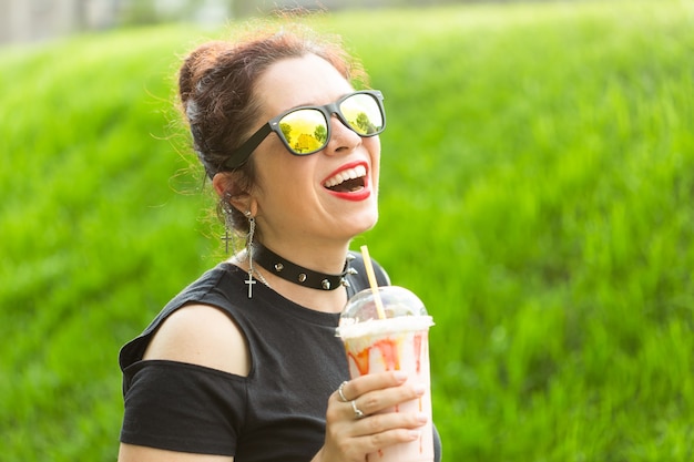 Ritratto di una giovane bella ragazza positiva in abiti punk e occhiali con un frappè in mano