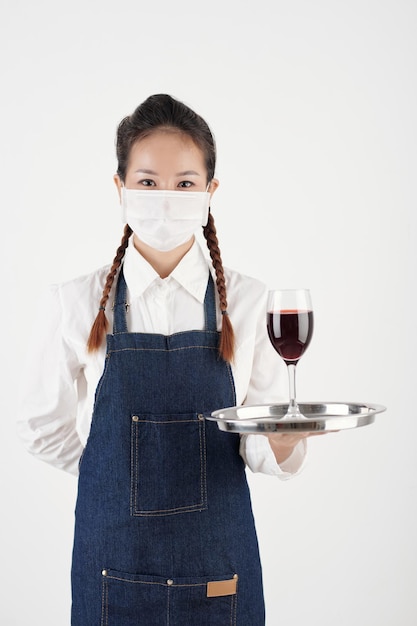 Ritratto di giovane cameriera positiva in maschera protettiva che tiene il vassoio con un bicchiere di vino rosso