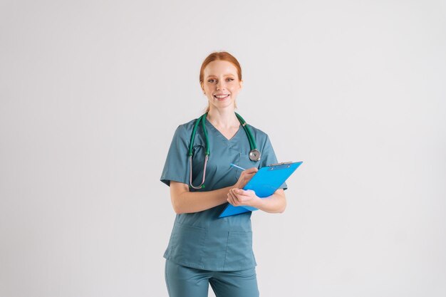 Portrait of positive young woman nurse in medical green uniform writing prescription on clipboard fills out medical history looking at camera standing on white isolated background in studio