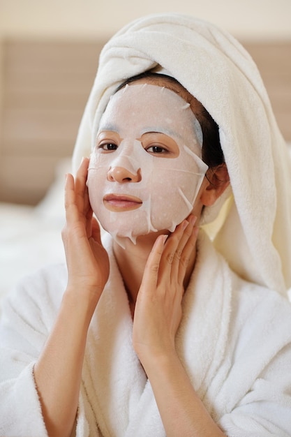 Portrait of positve young woman applying soothing and hydrating sheet mask on face after morning shower