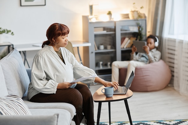 Portrait of pregnant African-American woman using laptop at home with teenage son in background, copy space