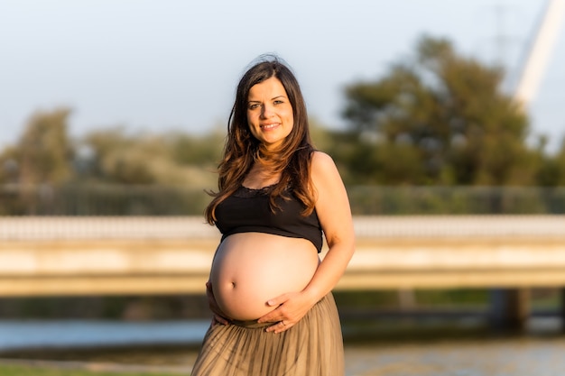 Portrait of a pregnant latina woman smiling to the camera while touching her belly standing on a urban lake