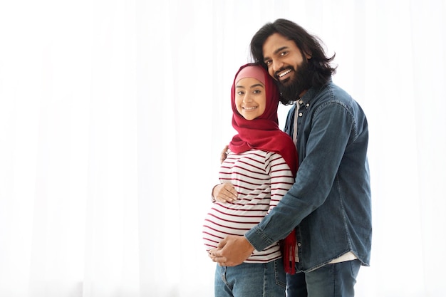 Photo portrait of pregnant muslim couple embracing near window at home
