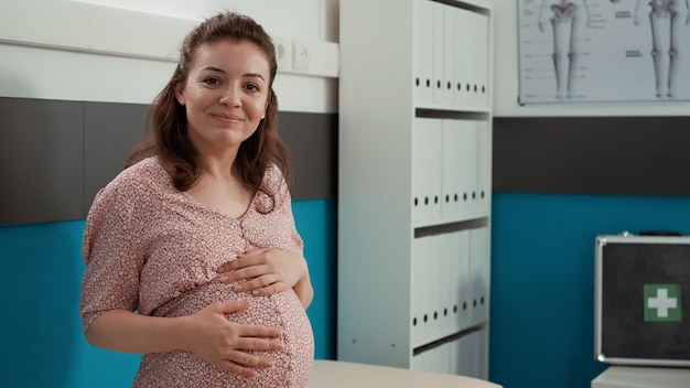 Portrait of pregnant patient waiting for doctor to attend physical examination in cabinet. Expectant woman with baby bump sitting on bed to meet with specialist at checkup consultation.