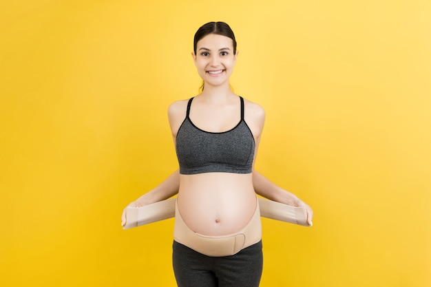 Portrait of pregnant woman dressing maternity belt against pain in the back at yellow background with copy space. Orthopedic abdominal support belt concept.