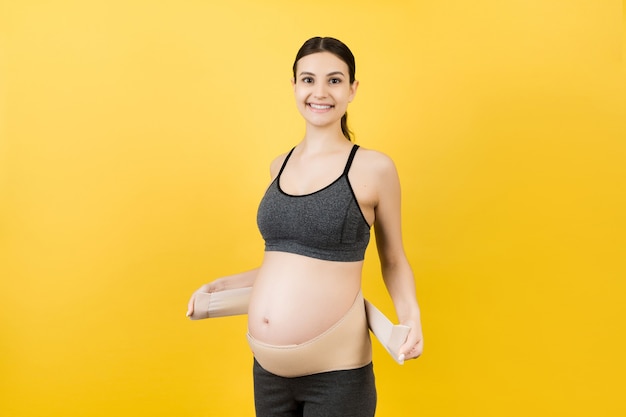 Portrait of pregnant woman dressing maternity belt against pain in the back at yellow background with copy space. Orthopedic abdominal support belt concept.