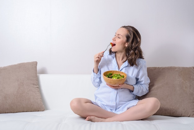 Портрет беременной женщины на диване, едят салат из свежих зеленых овощей