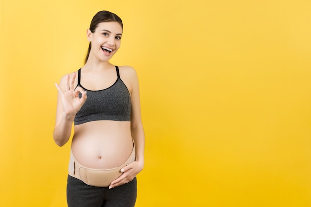 Portrait of pregnant woman on the third trimester wearing pregnancy belt and showing okay sign at yellow background. Orthopedic abdominal support belt concept with copy space.