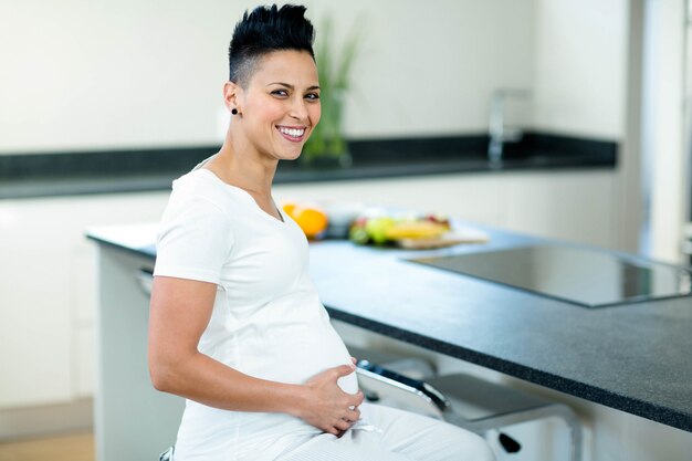 Portrait of pregnant woman touching her stomach while sitting near kitchen worktop