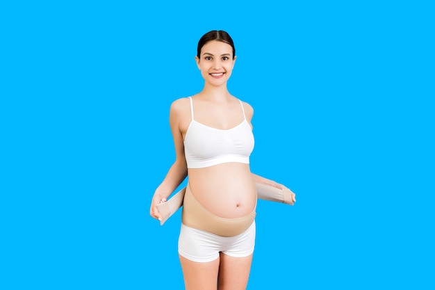 Portrait of pregnant woman in underwear putting on supporting bandage for reducing backache at blue surface with copy space