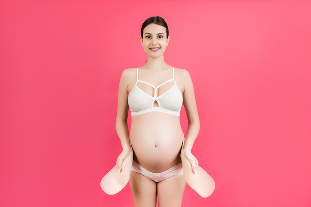 Портрет беременной женщины в нижнем белье, надевающей поддерживающую повязку для уменьшения боли в спине на розовом фоне с копией пространства. Ортопедическая концепция пояса поддержки живота.