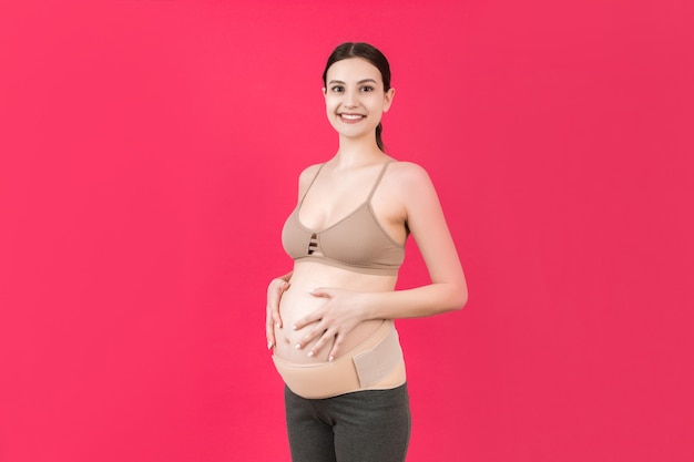 복사 공간이 있는 분홍색 배경에서 허리 통증을 줄이기 위해 출산 벨트를 착용한 임산부의 초상화. 정형 복부 지원 벨트 개념입니다.
