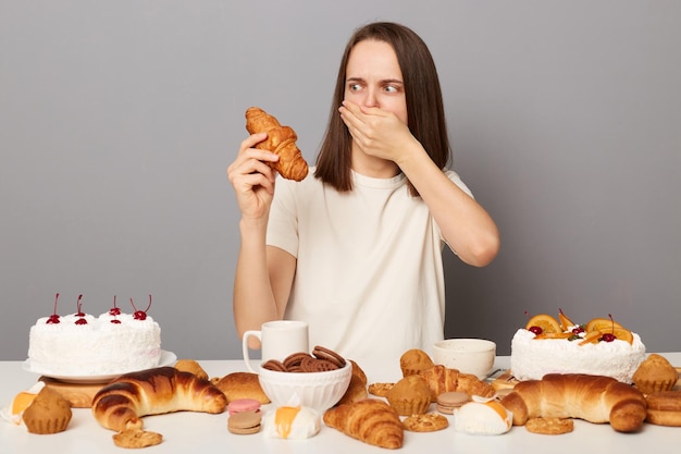 Portrait of pregnant woman with brown hair wearing white Tshirt sitting at table with different desserts feels nausea from aromat of croissant isolated over gray background