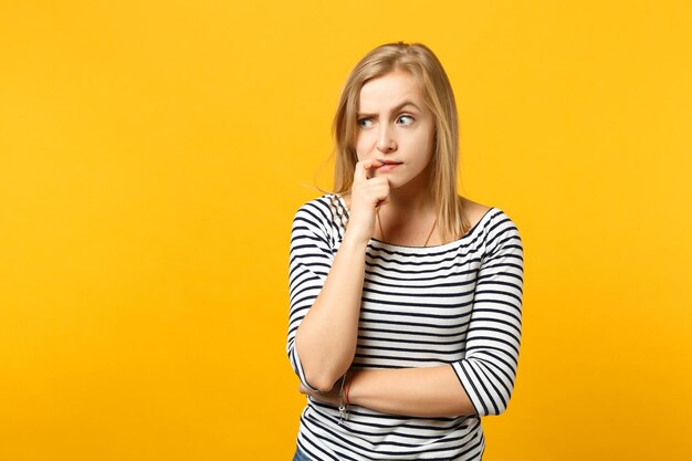 Portrait of preoccupied young woman in striped clothes looking aside, gnawing nails isolated on yellow orange wall background in studio. People sincere emotions, lifestyle concept. Mock up copy space.