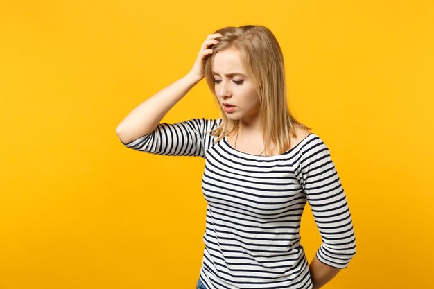 Portrait of preoccupied young woman in striped clothes looking down aside putting hand on head isolated on yellow orange wall background. People sincere emotions lifestyle concept. Mock up copy space.