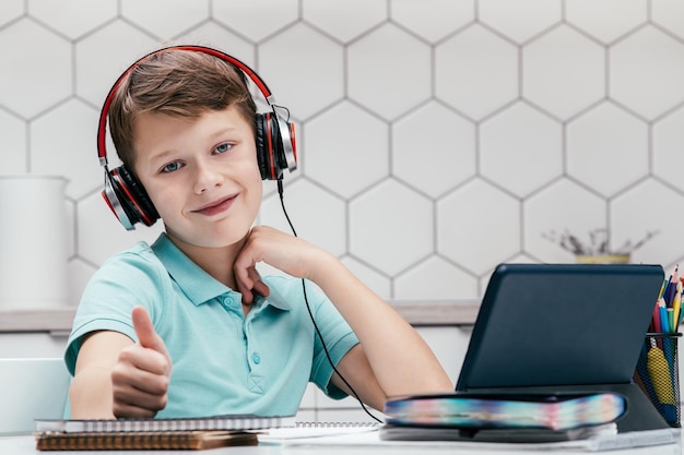Portrait of preteen boy sitting at desk in front of tablet near notebooks showing thumb up sign Approving technology