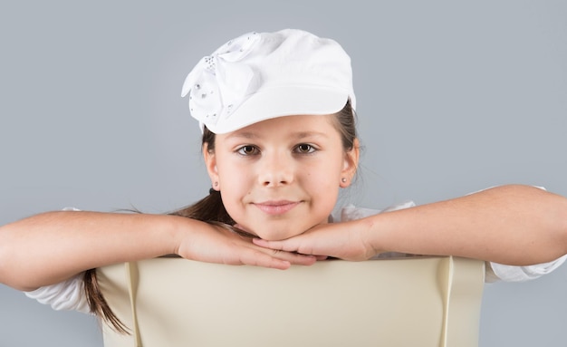 Portrait of a preteen girl in white cap sitting astride a chair and dreaming