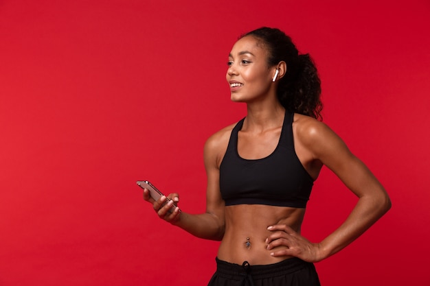 赤い壁に隔離されたスマートフォンとワイヤレスイヤホンを使用して黒いスポーツウェアでかなりアフリカ系アメリカ人の女性の肖像画