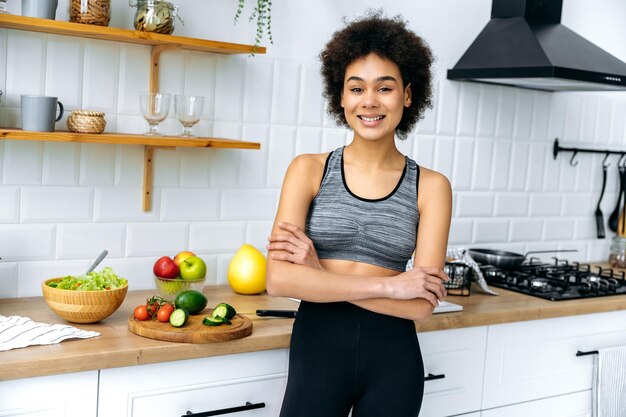 Foto ritratto di una bella giovane donna afroamericana riccia vestita in abbigliamento sportivo in piedi in cucina con le braccia incrociate guarda la telecamera sorride verdure fresche per l'insalata si trovano sullo sfondo