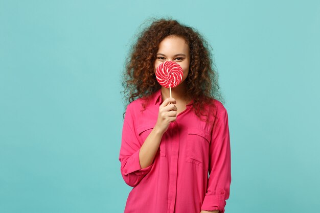 Photo portrait of pretty african girl in casual clothes covering face with pink round lollipop isolated on blue turquoise background in studio. people sincere emotions lifestyle concept. mock up copy space.