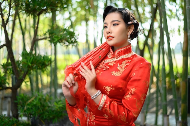 Portrait pretty Asian woman in a Chinese cheongsam posing with beautiful red paper umbrella on bamboo forest