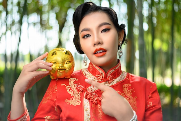 Portrait pretty Asian woman in a Chinese cheongsam posing with piggy bank on bamboo forest