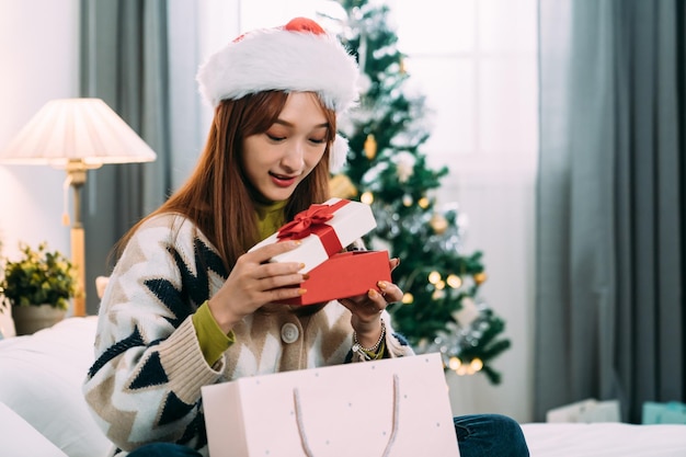 침대에 종이 봉지 옆에 앉아 있는 예쁜 아시아 여성이 집에 있는 축제 침실에서 박싱 데이에 크리스마스 선물을 여는 동안 상자 안을 들여다보고 있습니다.
