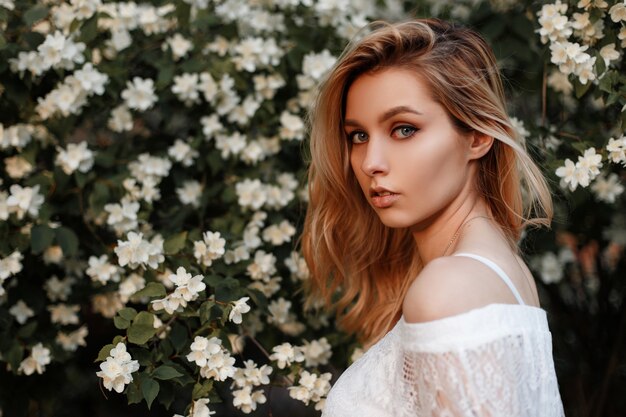 Portrait of a pretty attractive young blond woman with blue beautiful eyes with natural make-up in a vintage lace blouse near flowering bushes outdoors in spring day