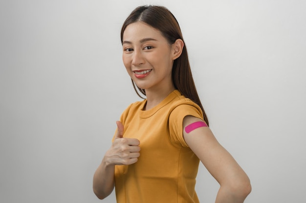 Portrait of pretty, beautiful asian young, teenage attractive after getting, receive anti virus vaccine covid-19. Showing arm on pink bandage in yellow t-shirt isolated on white background, copy space