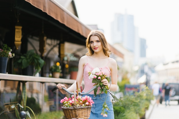 Portrait of a pretty and beautiful girl on the street of the city, bathed in the setting sun. The girl is holding a bouquet of roses and holding a Bicycle handlebar. The concept of summer walks