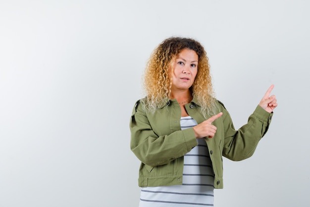 Portrait of pretty blond woman pointing at upper right corner in green jacket and looking hesitative front view