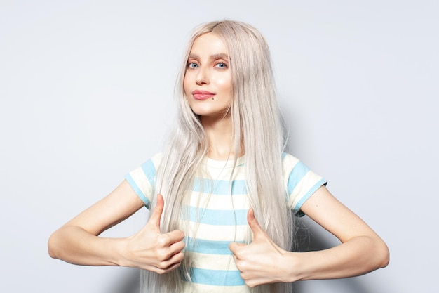 Портрет симпатичной блондинки в полосатой рубашке, показывающей большие пальцы на белом