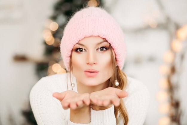 Portrait of pretty blonde woman in pearl earrings wearing pink winter woolen hat blowing air kiss at front with her hands, looking at front