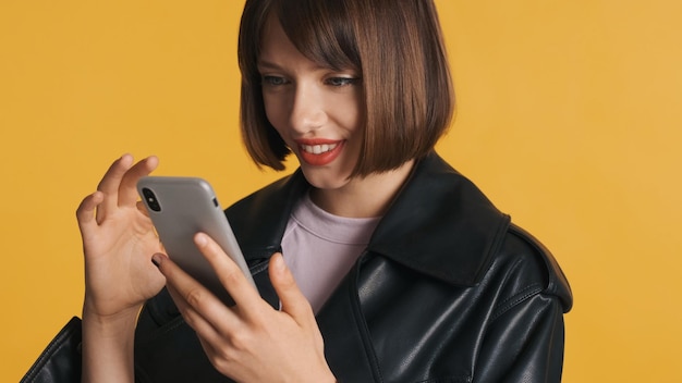 portrait of pretty brunette girl with bob hair in casual wear happily surfes social media on smartphone isolated on yellow background