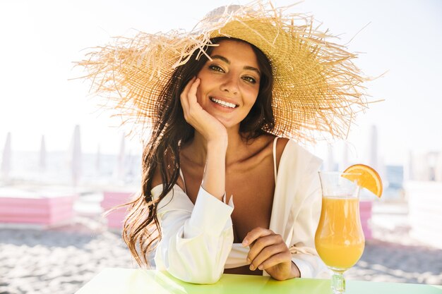 Portrait of pretty brunette woman in straw hat and summer clothes drinking orange juice in beach cafe on vacation