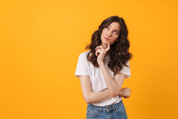 Portrait of pretty brunette woman wearing casual clothes thinking and looking upward at copyspace isolated over yellow wall