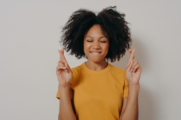 Portrait of pretty cheerful african girl standing isolated over white studio background with copy space, holding fingers crossed for good luck, with closed eyes biting lower lip while making wish