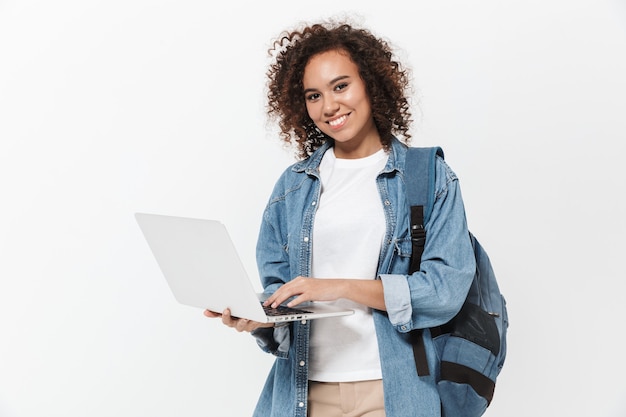 Portrait of a pretty cheerful casual african girl carrying backpack standing isolated over white wall, holding laptop computer