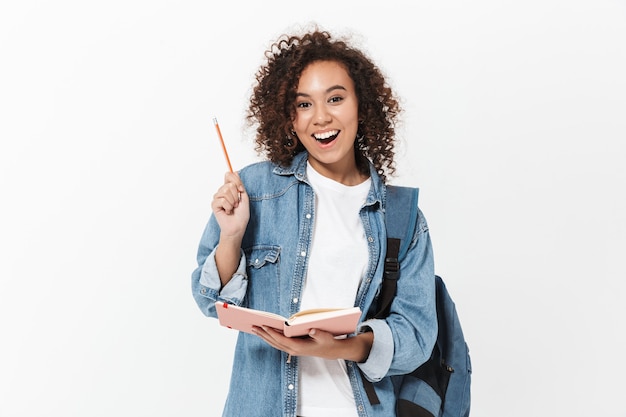 Portrait of a pretty cheerful casual african girl carrying backpack standing isolated over white wall, writing in a textbook