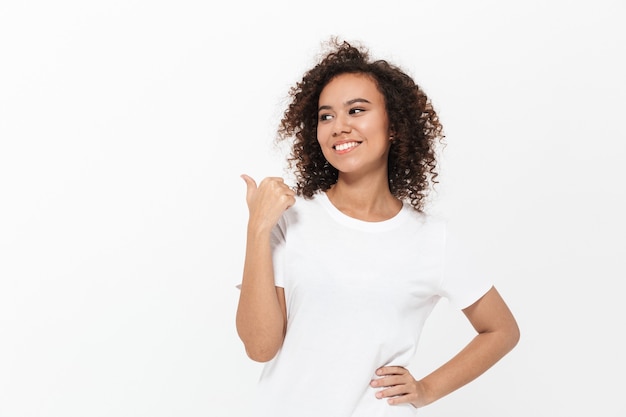 Portrait of a pretty cheerful casual african girl standing isolated over white wall, presenting copy space