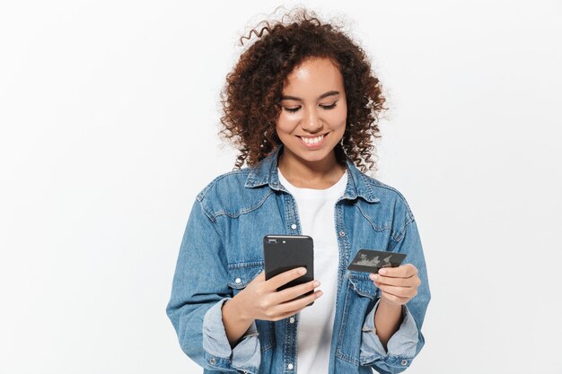 Ritratto di una ragazza africana casual piuttosto allegra in piedi isolata su un muro bianco, mostrando una carta di credito in plastica, usando il telefono cellulare